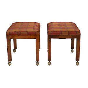 May include: Two wooden ottomans with plaid cushions in shades of red, orange, and brown. The ottomans have square seats, four legs, and brass caster wheels. The wood has a warm, reddish-brown tone.