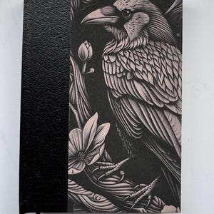 Puede incluir: Un diario negro con una ilustración detallada de un cuervo y flores en la portada. El cuervo está representado en tonos rosas y blancos sobre un fondo negro. La columna vertebral es negra y texturizada.