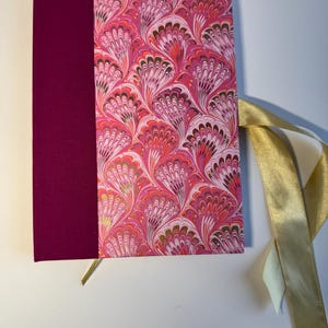 Könnte beinhalten: Ein Hardcover-Journal mit einem lebendigen rosa-weißen Marmormuster auf dem Einband. Der Buchrücken ist einfarbig magenta. Ein goldenes Leseband und ein goldenes Satinband sind befestigt.