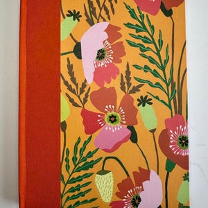 May include: A hardback journal featuring a vibrant mustard yellow cover with a repeating pattern of red and pink poppies, green foliage, and light green seed pods. The spine is bound in a solid burnt orange fabric with a matching ribbon bookmark.