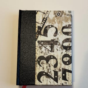Puede incluir: Un pequeño diario rectangular con lomo negro texturizado y cubierta estampada. La cubierta presenta grandes números negros e imágenes de carretes de película sobre un fondo blanquecino desgastado. Una cinta roja como marcapáginas es visible.