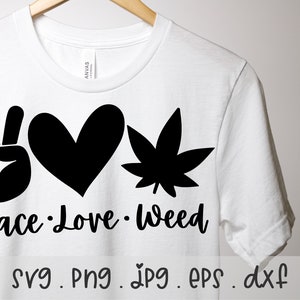 May include: White t-shirt with a black graphic featuring a peace sign, a heart, and a cannabis leaf. The words "Peace Love Weed" are printed below the symbols. The shirt is hanging on a wooden hanger.