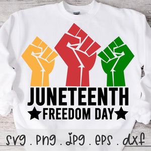 May include: White sweatshirt featuring three raised fists in red, yellow, and green. The text "Juneteenth Freedom Day" is printed in black below the fists.