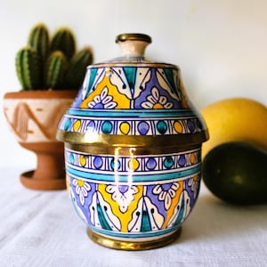 Op de afbeelding: Een decoratieve keramische pot met deksel, met een kleurrijk bloemmotief in blauw, geel en teal. De pot heeft goudkleurige metalen accenten en staat op een wit oppervlak. Een cactus in een terracotta pot, een citroen en een avocado staan op de achtergrond.