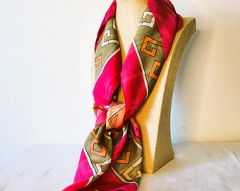 May include: A vibrant pink silk scarf with a geometric pattern in olive green, white, and orange. The scarf is draped over a mannequin head, showcasing its design and texture. The scarf is knotted at the bottom.