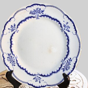 May include: A white ceramic plate with a blue and white floral design. The plate has a scalloped edge and is decorated with a repeating pattern of flowers and leaves.