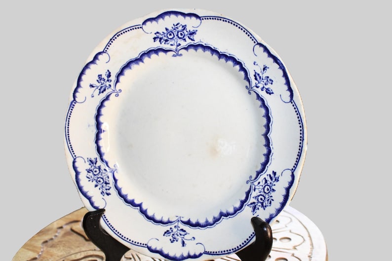 May include: A white ceramic plate with a blue and white floral design. The plate has a scalloped edge and is decorated with a repeating pattern of flowers and leaves.