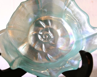 May include: A decorative, iridescent glass bowl with a seashell design. The bowl has a light blue hue with a pearlescent sheen. The bowl is supported by a black stand. The bowl's dimensions are approximately 8 inches in diameter.