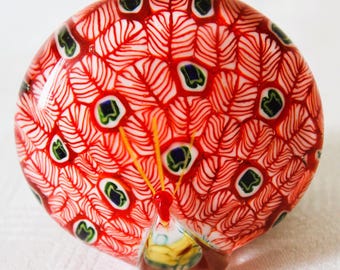 May include: A decorative glass peacock figurine. The figurine features a red and white pattern resembling feathers, with accents of green, blue, and yellow. The peacock's tail is fanned out, and the base is brown.