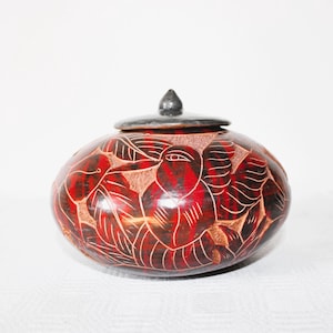 Puede incluir: Un tarro decorativo rojo y marrón con tapa. El tarro presenta un diseño tallado de pájaros y hojas. La tapa es de color gris oscuro y tiene un pequeño pomo puntiagudo. El tarro está sobre una superficie blanca.