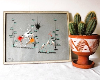 May include: An abstract artwork in a white framed with a grey background, featuring colourful shapes and figures. A terracotta pot with a cactus is to the right. The pot has a white and orange wave pattern.