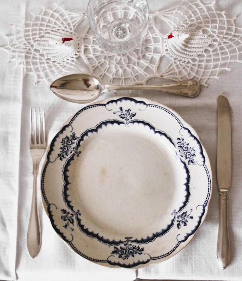 May include: A white porcelain dinner plate with a blue and white floral pattern. The plate is set on a white tablecloth with a silver fork, spoon, and knife.