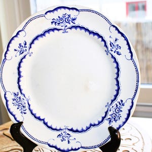 May include: A white ceramic plate with a blue and white floral design. The plate has a scalloped edge and is decorated with a repeating pattern of flowers and leaves.