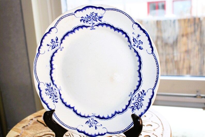 May include: A white ceramic plate with a blue and white floral design. The plate has a scalloped edge and is decorated with a repeating pattern of flowers and leaves.