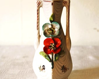May include: A white ceramic vase with a square base and two handles. The vase is decorated with a red and blue flower design with green leaves. The vase has a textured wood grain pattern and small floral accents.
