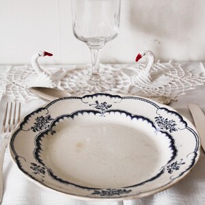 May include: A white porcelain dinner plate with a blue and white floral pattern. The plate is set on a white tablecloth with a white crocheted doily and two white swan figurines.