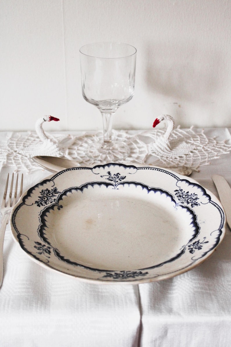 May include: A white porcelain dinner plate with a blue and white floral pattern. The plate is set on a white tablecloth with a white crocheted doily and two white swan figurines.