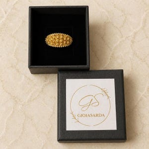 May include: A gold-colored filigree ring in a black jewelry box. The ring has a detailed, textured design. The open box below displays a logo with the text "GIOIASARDA".