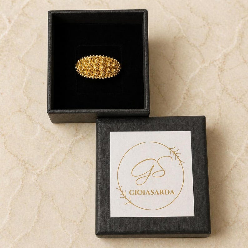 May include: A gold-colored filigree ring in a black jewelry box. The ring has a detailed, textured design. The open box below displays a logo with the text "GIOIASARDA".