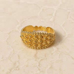 May include: A gold-colored ring with a detailed, textured design. The ring features a pattern of small, raised dots and floral-like shapes, creating a delicate and ornate appearance. The ring is likely made of gold and is a piece of jewelry.