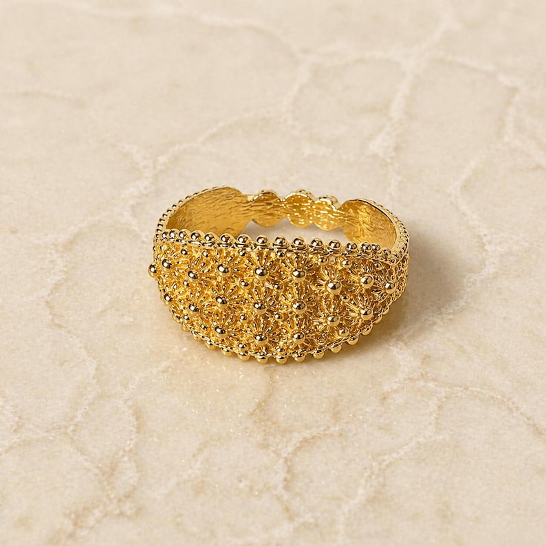 May include: A gold-colored ring with a detailed, textured design. The ring features a pattern of small, raised dots and floral-like shapes, creating a delicate and ornate appearance. The ring is likely made of gold and is a piece of jewelry.