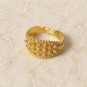 May include: A gold-colored ring with an intricate, textured design. The ring features a detailed pattern of small, raised dots and floral-like shapes, creating a delicate and ornate appearance. The ring is set against a neutral background.