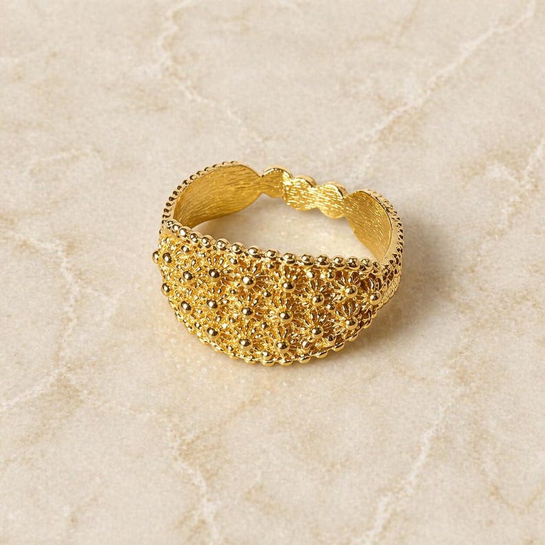 May include: A gold-colored ring with an intricate, textured design. The ring features a detailed pattern of small, raised dots and floral-like shapes, creating a delicate and ornate appearance. The ring is set against a neutral background.
