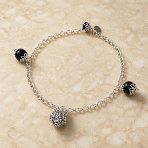 Puede incluir: Una pulsera plateada con un diseño de cadena, con cuatro elementos decorativos. La pulsera incluye tres cuentas negras esféricas con tapas plateadas ornamentadas y una cuenta plateada con un patrón detallado. La pulsera se muestra sobre una superficie clara.