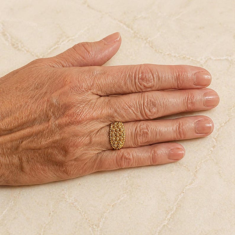 May include: A gold-colored ring with an oval design, featuring a textured, woven pattern. The ring is worn on a finger, showcasing its intricate details and craftsmanship. The ring's warm hue contrasts with the skin tone, highlighting its elegance.