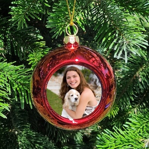 May include: Red Christmas bauble with a photograph of a woman and a dog inside. The woman is smiling and holding the dog. The dog is a brown and white Cocker Spaniel.