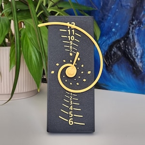May include: A rectangular black clock with a textured surface featuring a gold spiral design. The clock face displays vertical numerical markings from 1 to 12 in gold, arranged in a linear sequence with a central gold hand pointing toward the centre.