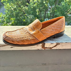 May include: A single tan leather loafer with a woven design on the vamp and sides. The shoe has a leather strap across the top and a black sole. The shoe is sitting on a wooden surface.