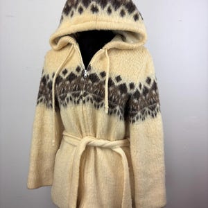 May include: A cream-colored hooded sweater with a brown and white geometric pattern. The sweater has a zipper closure and a belt.