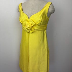 May include: A bright yellow sleeveless dress with a V-neckline and a decorative flower detail at the waist. The dress is displayed on a mannequin and has a fitted silhouette. The fabric appears to be a smooth, possibly satin, material.