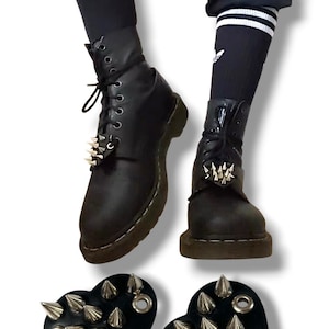 May include: Black leather boots with silver spike details and black socks with white stripes. Two black heart-shaped accessories with silver spikes are also shown. The boots have a lace-up design.