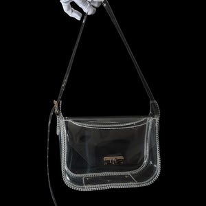May include: A clear, transparent crossbody bag with a silver-colored chain trim and strap. The bag has a rectangular shape with a curved bottom and a clasp closure. The background is black.