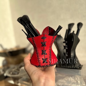 Könnte beinhalten: Roter Leder-Make-up-Pinselhalter in Form eines Korsetts mit schwarzen Nähten und schwarzen Schnüren. Der Halter ist mit schwarzen Make-up-Pinseln gefüllt. Das Wort "MIRAMUR" ist auf dem schwarzen Halter im Hintergrund sichtbar.
