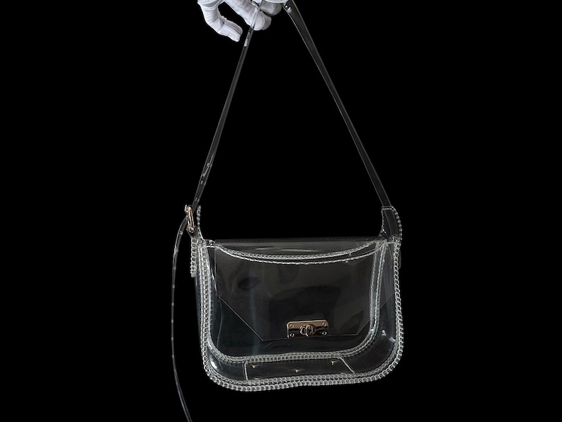 May include: A clear, transparent crossbody bag with a silver-colored chain trim and strap. The bag has a rectangular shape with a curved bottom and a clasp closure. The background is black.
