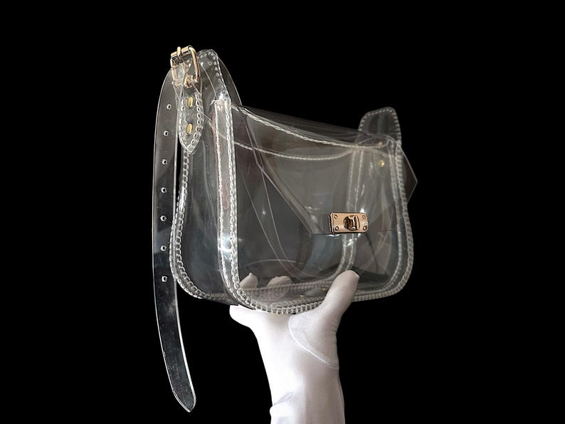 May include: Clear, transparent crossbody bag with a gold-colored clasp and hardware. The bag has a flap closure and a long, adjustable strap. The bag is held up by a gloved hand against a black background.