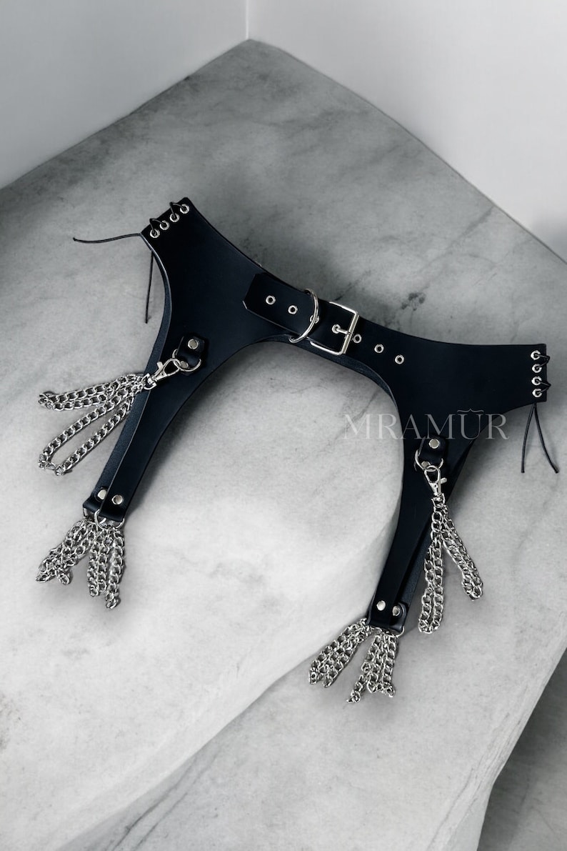 May include: Black leather garter belt with silver chain tassels. The belt has a buckle closure and multiple rows of eyelets. The text "MRAMUR" is visible on the belt.
