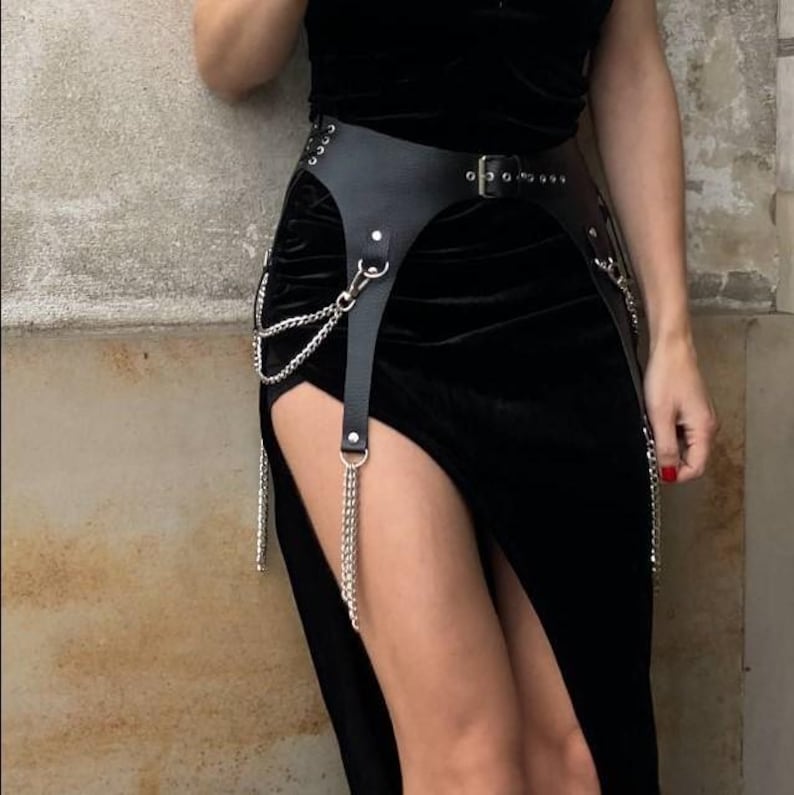 May include: Black leather harness with silver chain accents. The harness has a belt with a buckle and two straps that extend down the legs.