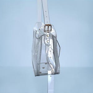 May include: A clear, transparent handbag with a shoulder strap and a gold-colored buckle. The bag is rectangular with rounded edges and features visible stitching and rivets. The strap is also clear and attached to the bag with a gold-colored buckle.