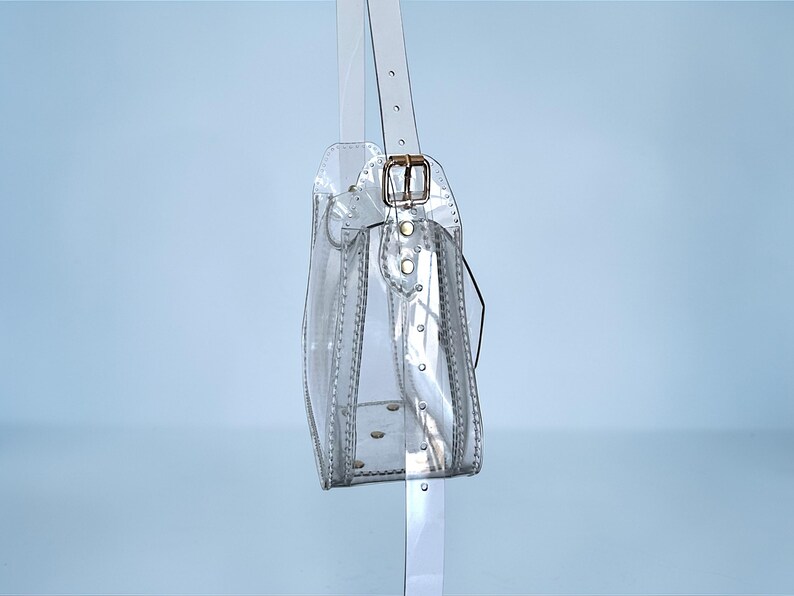May include: A clear, transparent handbag with a shoulder strap and a gold-colored buckle. The bag is rectangular with rounded edges and features visible stitching and rivets. The strap is also clear and attached to the bag with a gold-colored buckle.