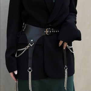 May include: A black blazer with a green satin slip dress underneath. A black leather belt with silver chains and a silver buckle is worn over the dress.