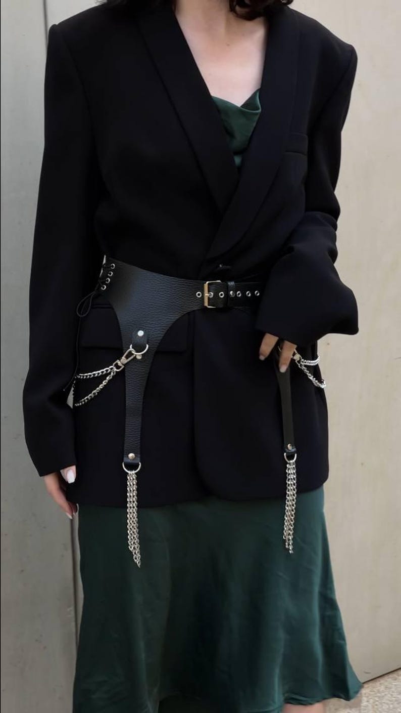May include: A black blazer with a green satin slip dress underneath. A black leather belt with silver chains and a silver buckle is worn over the dress.