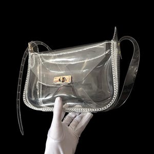 May include: Clear, transparent crossbody bag with a silver chain trim and a gold-colored clasp. The bag has a long, clear strap and is held against a black background.