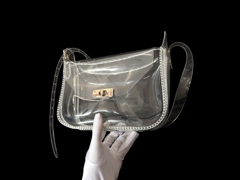 May include: Clear, transparent crossbody bag with a silver chain trim and a gold-colored clasp. The bag has a long, clear strap and is held against a black background.