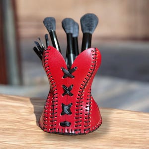 Puede incluir: Un soporte para brochas de maquillaje con forma de corsé de cuero rojo con costuras negras y detalles de lazo negro. El soporte contiene varios pinceles de maquillaje con mango negro y cerdas grises. El soporte está sobre una superficie de madera.
