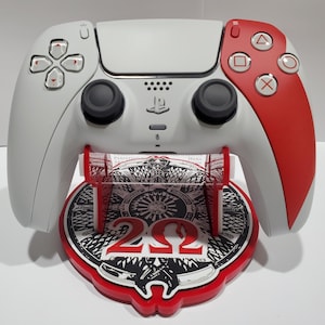 May include: A white and red PlayStation 5 controller resting on a matching stand. The controller has black joysticks and red action buttons. The stand is red with a black and white graphic design and the number "20".
