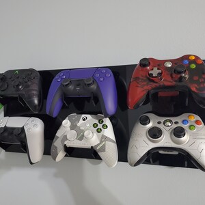 Pode incluir: Seis controladores de videogame diferentes montados em um suporte de parede preto. Os controladores s&atilde;o uma mistura de preto, branco, verde, vermelho e roxo. Os controladores s&atilde;o todos de marcas e modelos diferentes.